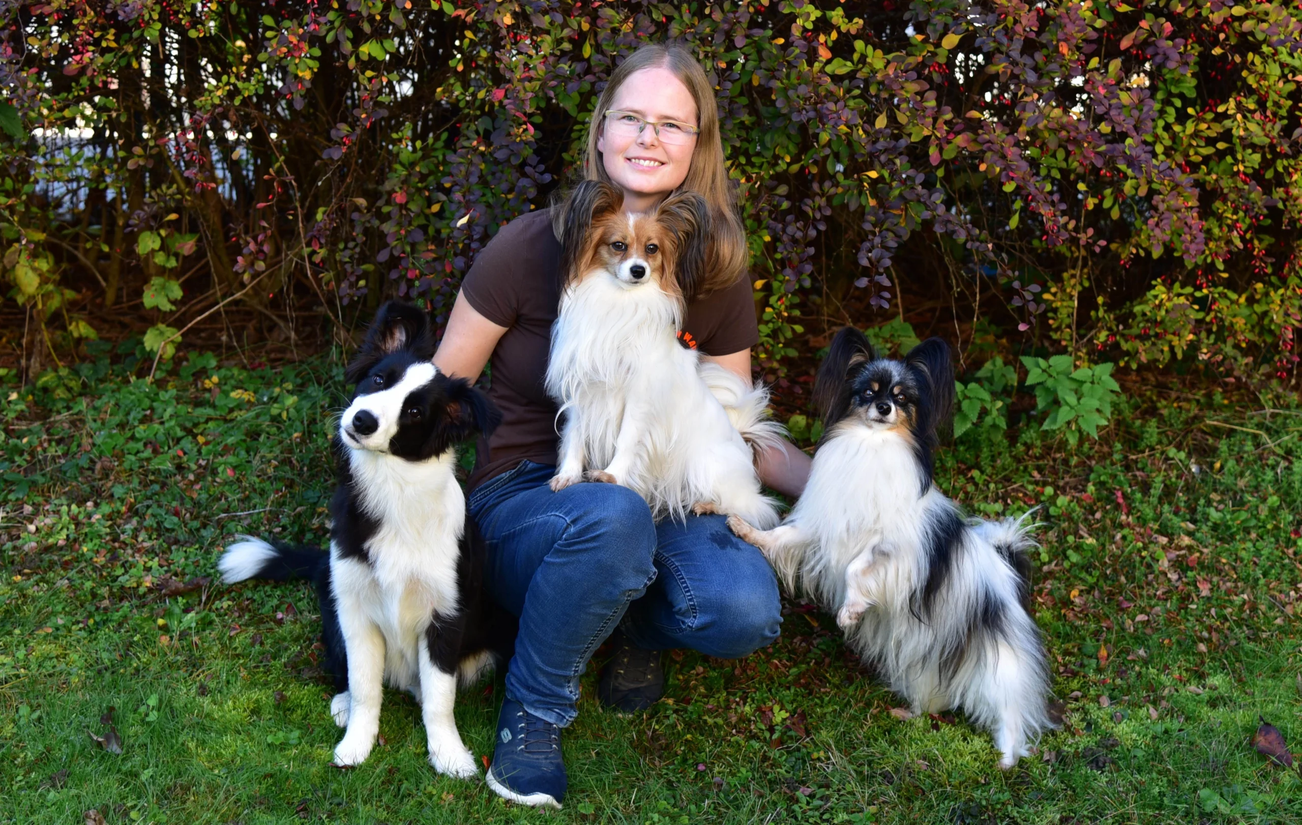 Lächelnde Frau kniet im Garten mit einem Hund auf ihrem Schoß und zwei Hunden neben ihr.