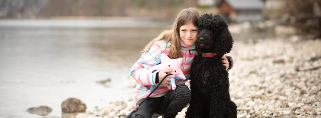 Mädchen kniet am Steinstrand und drückt Hund an sich.