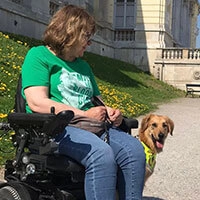 Assistenzhund sitzt hinter Frau in Rollstuhl.