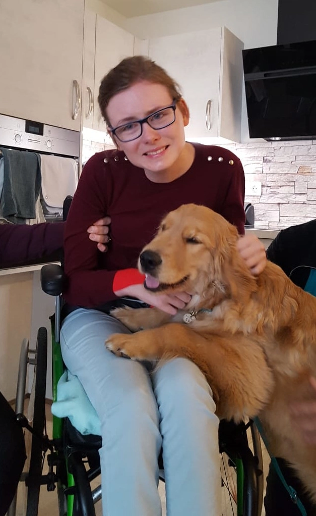 Lächelnde Frau in Rollstuhl mit ihrem Assistenzhund am Schoß.