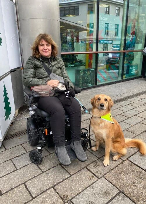 Eine Frau im Rollstuhl. Neben ihr sitzt ein Assistenzhund.