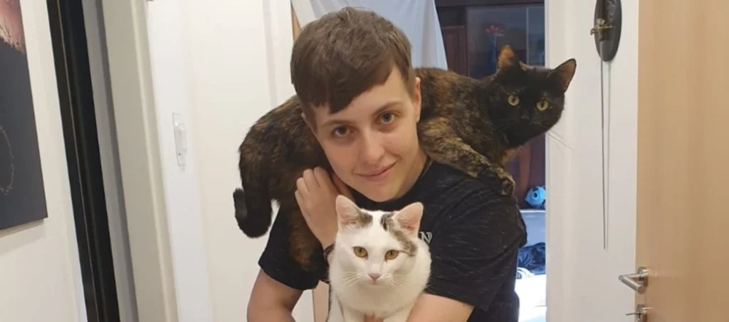 Person lächelt. Sie hält eine Katze in den Armen. Eine andere liegt auf ihren Schultern.