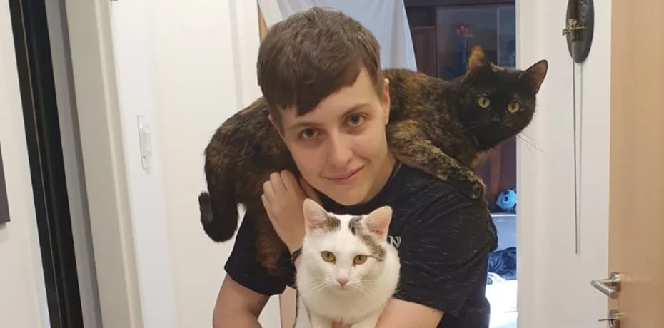 Person lächelt. Sie hält eine Katze in den Armen. Eine andere liegt auf ihren Schultern.