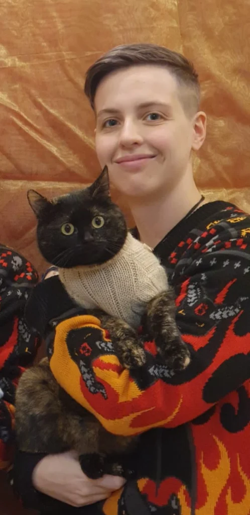 Person lächelt und hält eine Katze in den Armen. Die Katze hat einen ärmellosen Strickpullover an.